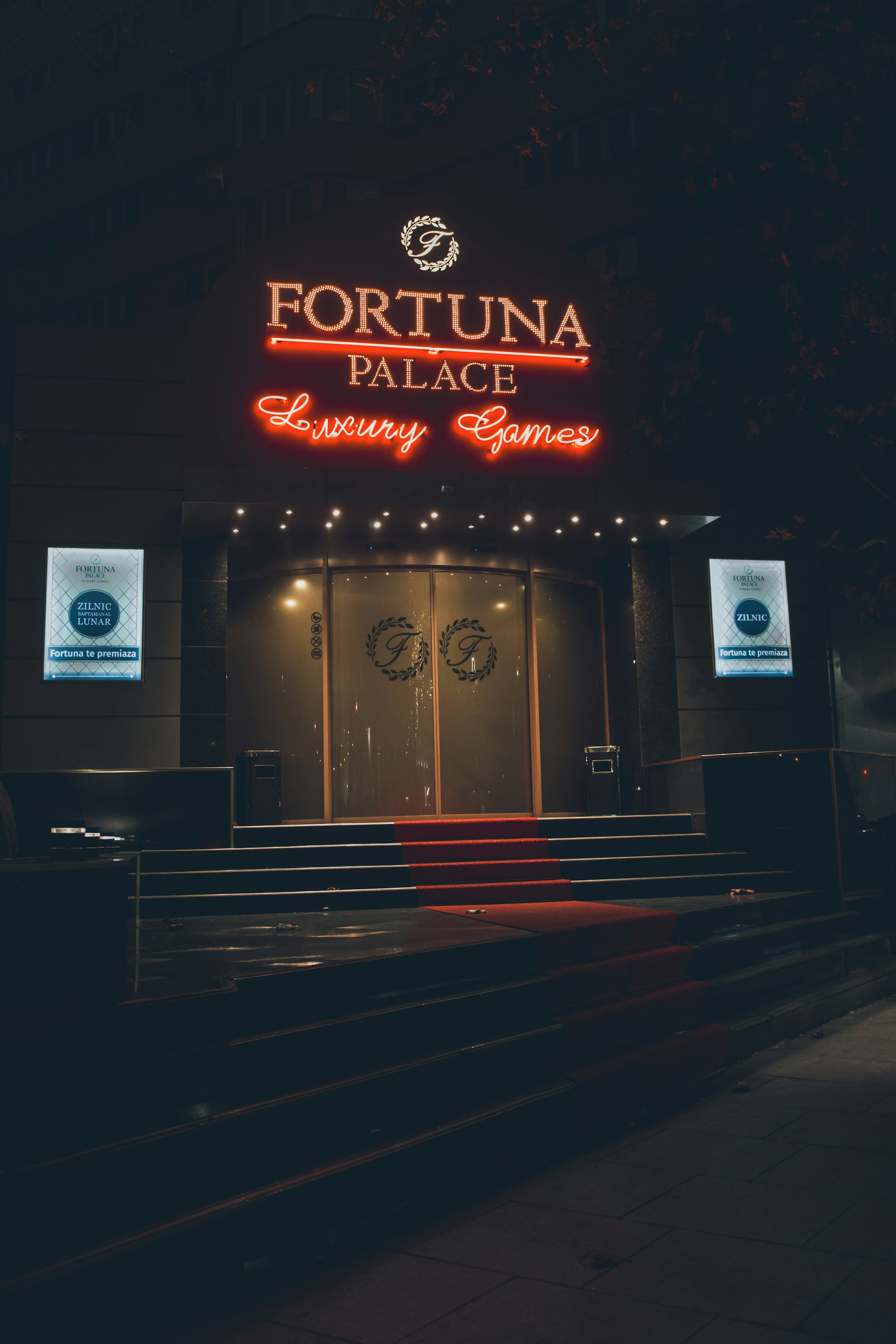 A neon-lit casino entrance with a red carpet leading up to large double doors. The sign reads 'Fortuna Palace Luxury Games' in bright red and white neon lights. The dimly lit atmosphere suggests a night-time setting. There are two digital displays on either side of the entrance showing promotional content.